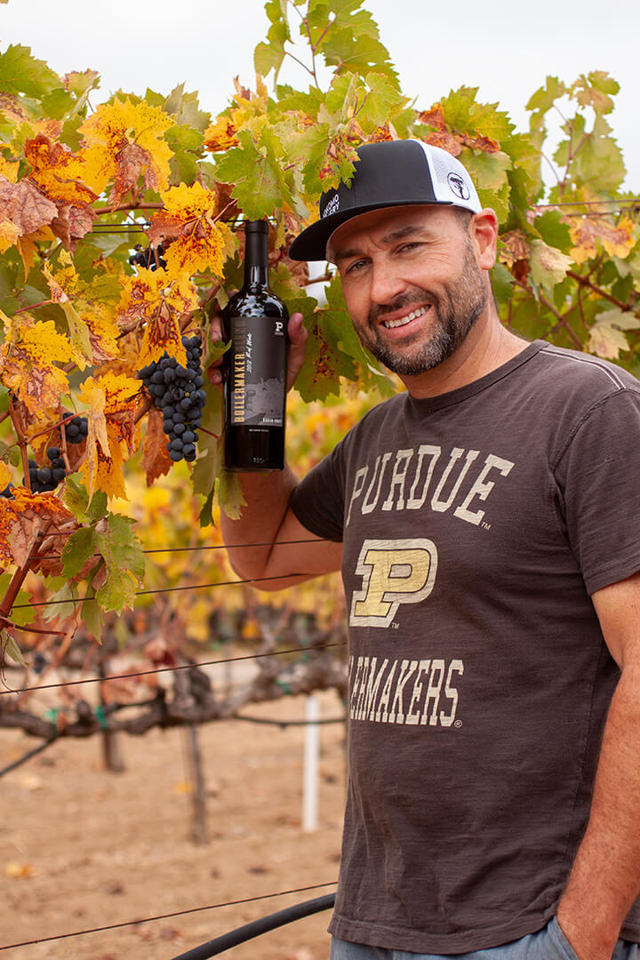 Eric Miller standing in front of vines holding bottle of boilermaker wine.